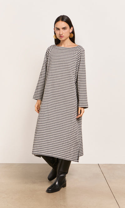 Plümo  Woman in Marsell Dress - Gingham, loose fit, slash neck, with black boots indoors against a plain wall.