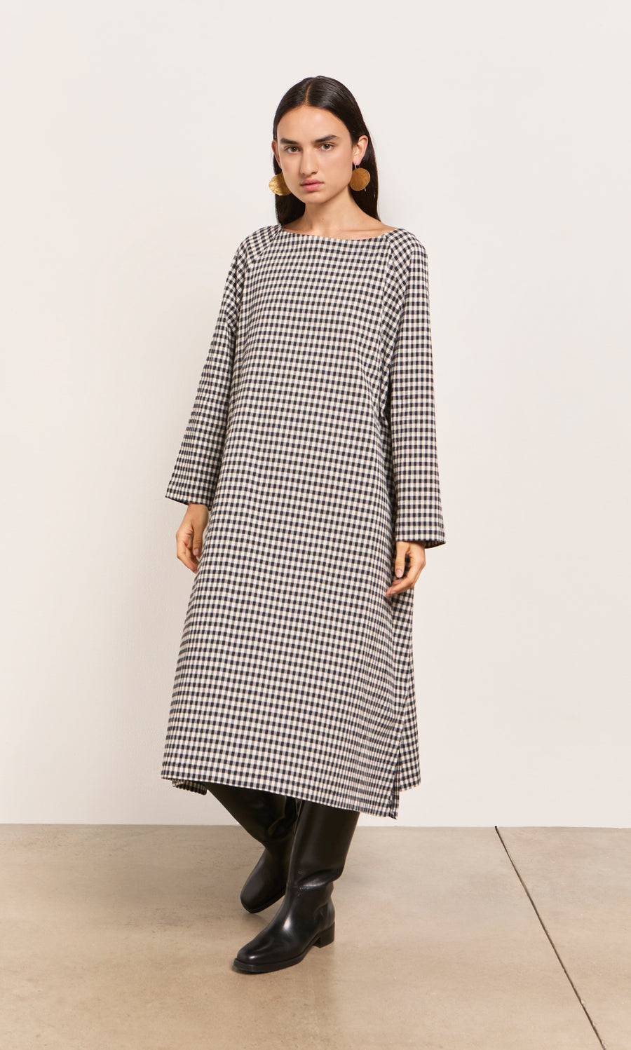 Plümo  Woman in Marsell Dress - Gingham, loose fit, slash neck, with black boots indoors against a plain wall.