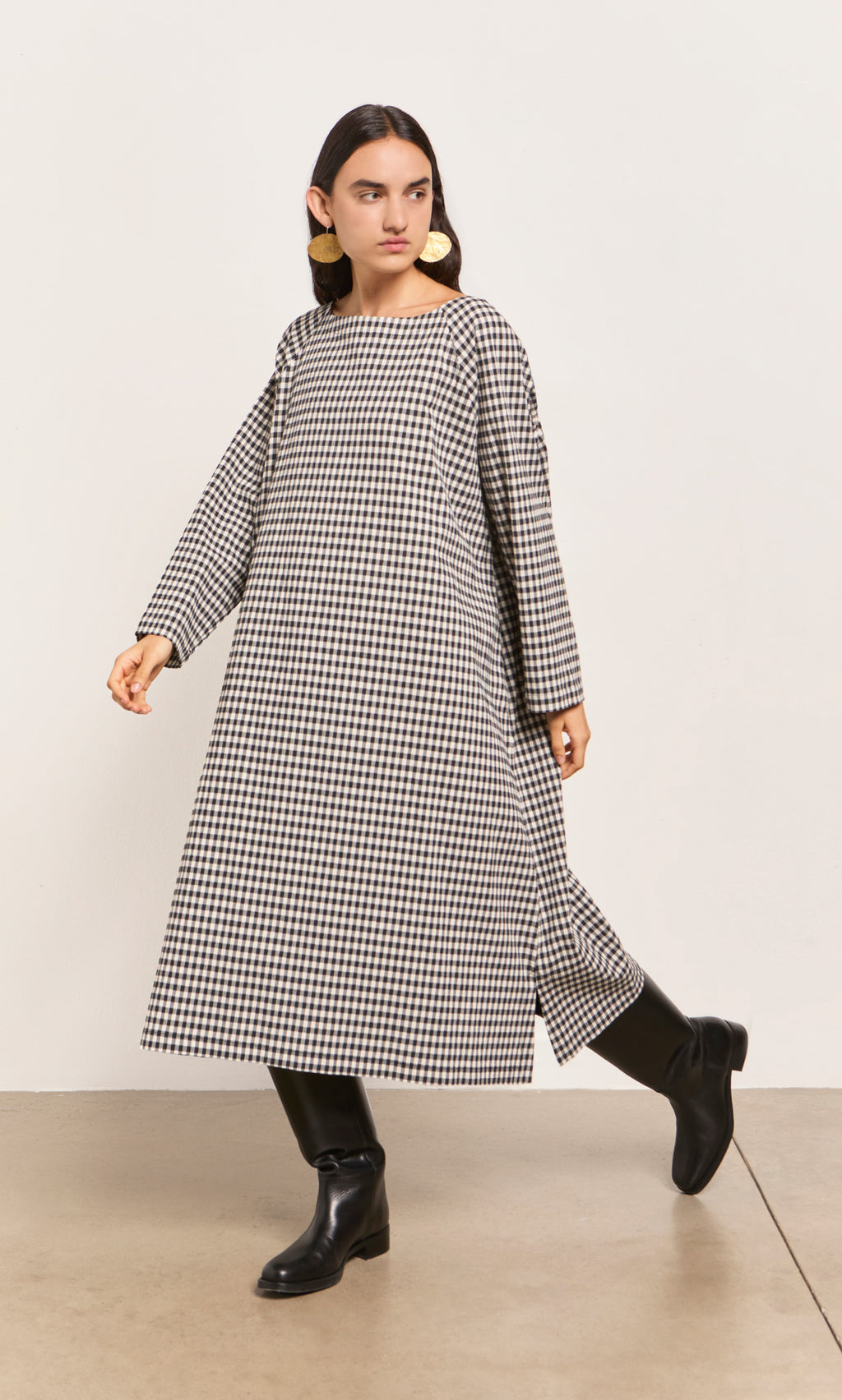 Plümo  Woman in the Marsell Dress - Gingham, black boots, and bold earrings walks indoors.