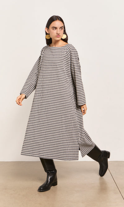 Plümo  Woman in the Marsell Dress - Gingham, black boots, and bold earrings walks indoors.