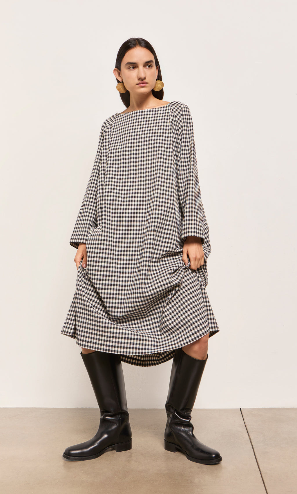 Plümo  Woman in a slash neck Marsell Gingham Dress and black knee-high boots stands against a plain background.