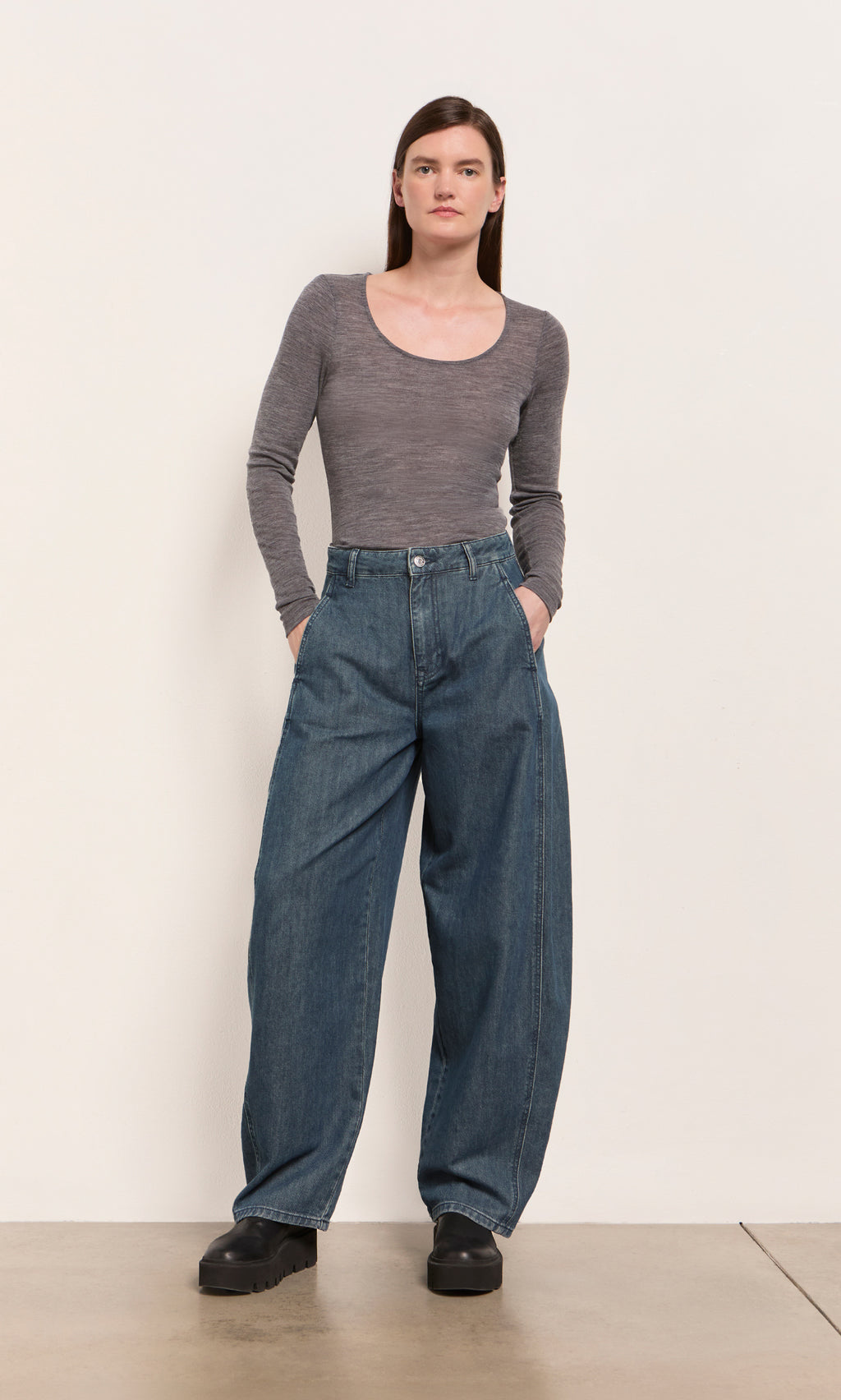 Plümo  Woman in a gray top and Maud Barrel Pants stands against a plain wall with hands in pockets.