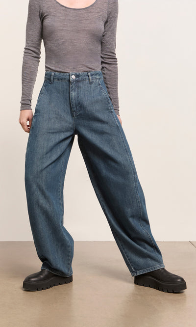 Plümo  Person wears Maud Barrel Pants, a gray long-sleeve shirt, and black boots while standing indoors.