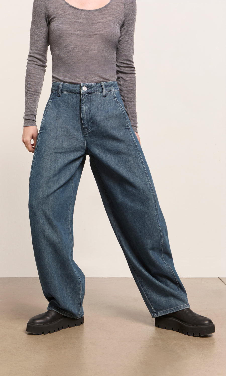 Plümo  Person wears Maud Barrel Pants, a gray long-sleeve shirt, and black boots while standing indoors.
