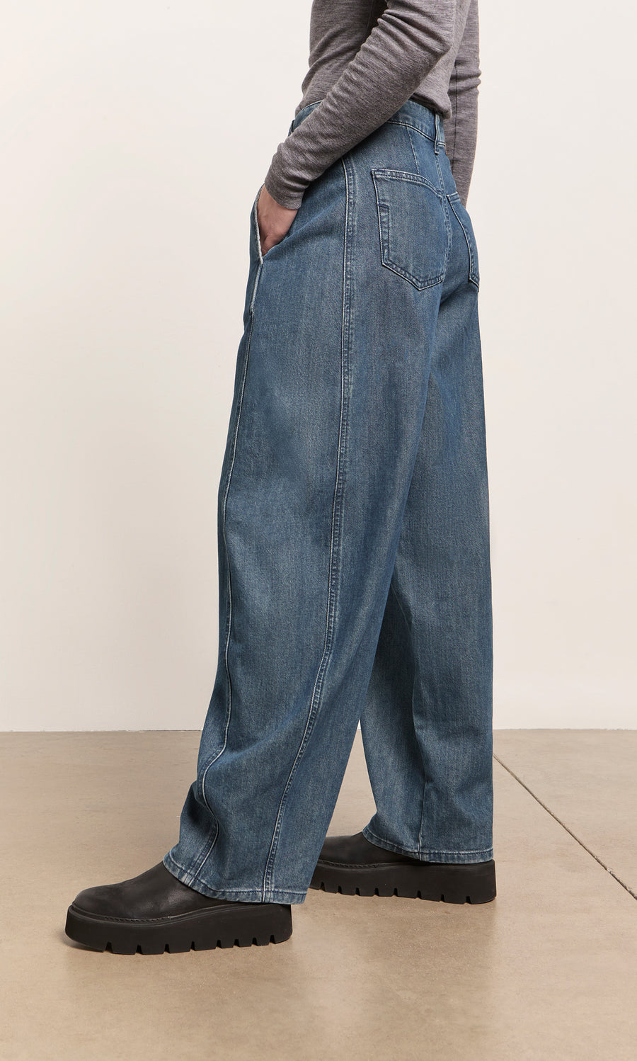 Plümo  Individual wears Maud Barrel Pants, a gray long-sleeve shirt, and black shoes, standing hands in pockets.