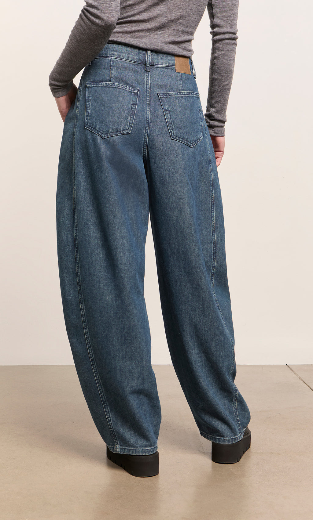 Plümo  Model wears Maud Barrel Pants in indigo wash and a gray long-sleeve shirt, standing on a beige floor facing away.