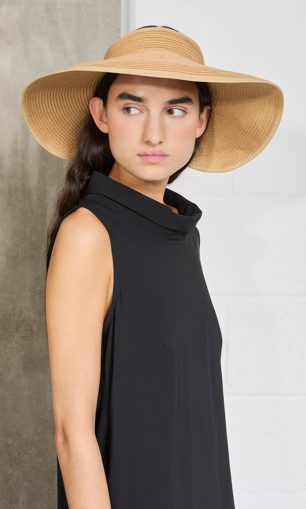 Plümo- Woman wearing the Tegan Dress, a comfortable fit sleeveless black dress, with a straw hat against a neutral background.
