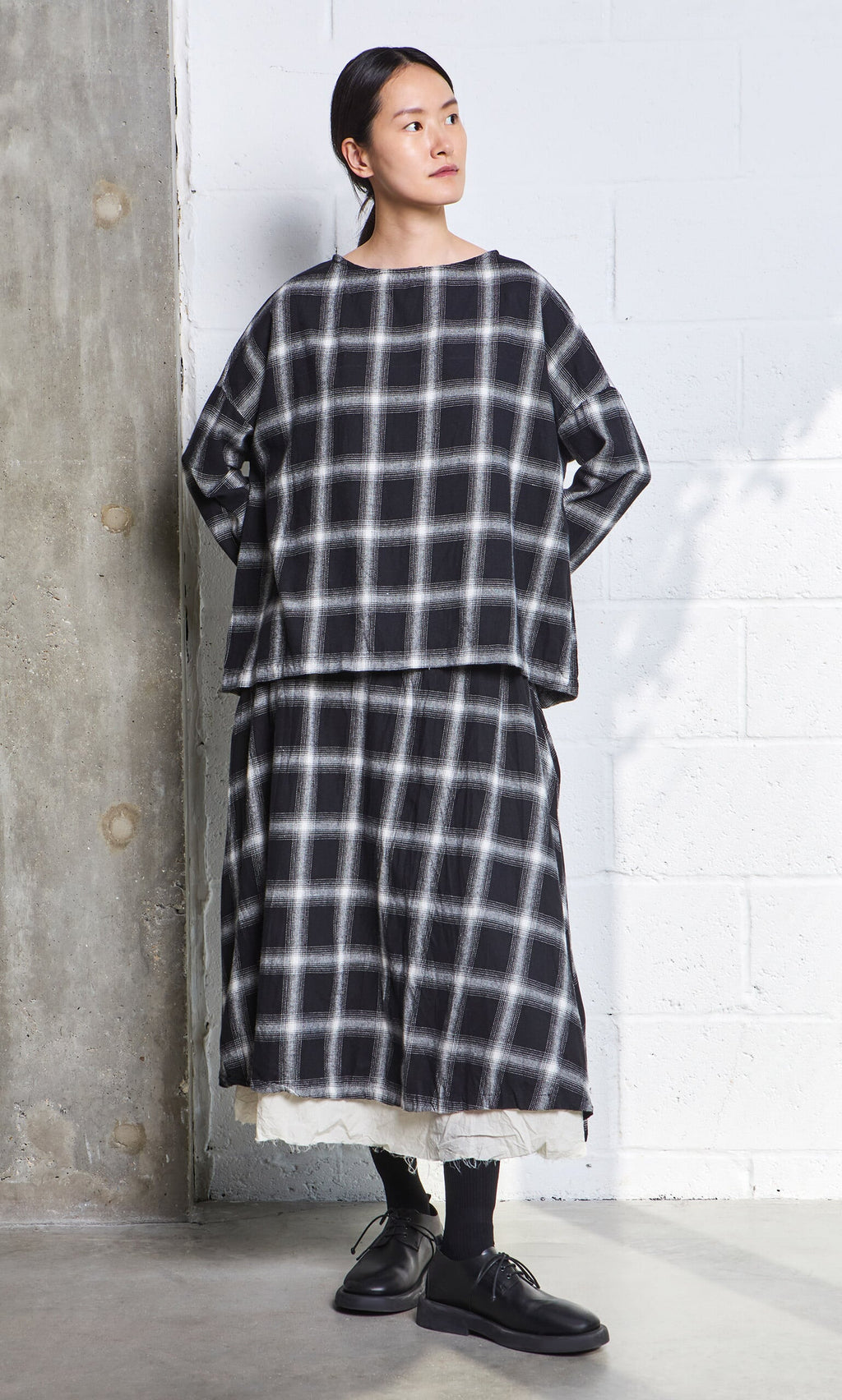 Plümo- Person wearing a Gifford Blouse and matching skirt in brushed flannel, with black socks and shoes by a concrete wall.
