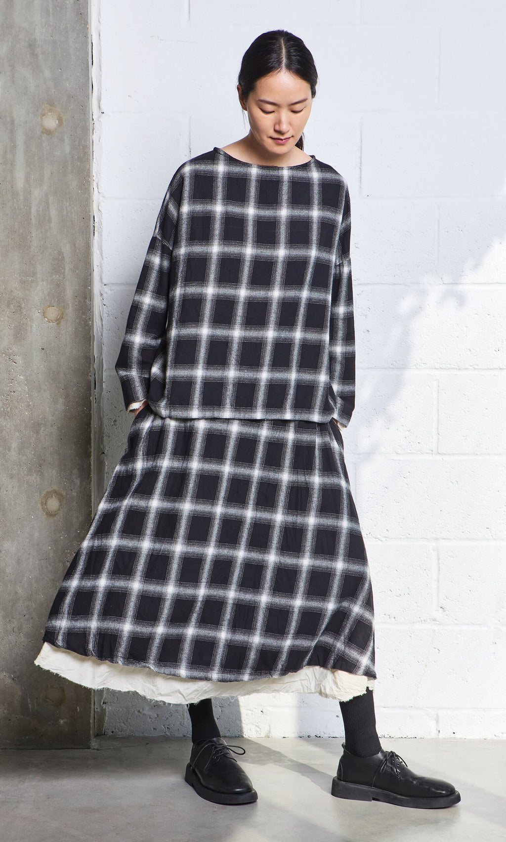 Plümo- Person wearing a loose, black and white plaid Noemi Skirt stands against a white and concrete wall.