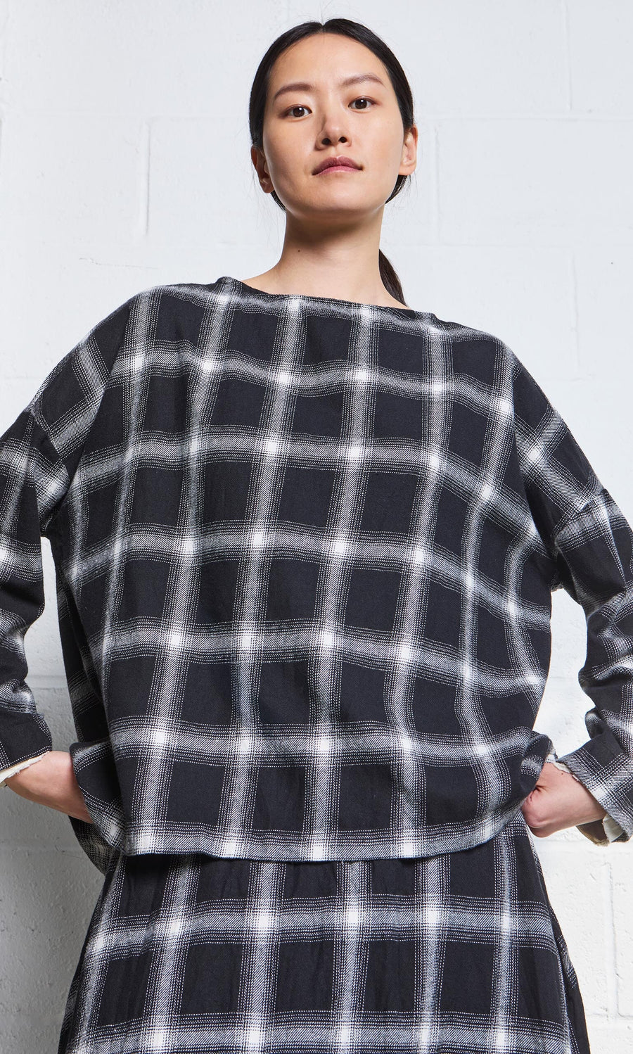 Plümo- Woman in the Italian-made Gifford Blouse, featuring a monochrome plaid design, stands confidently by a white wall.