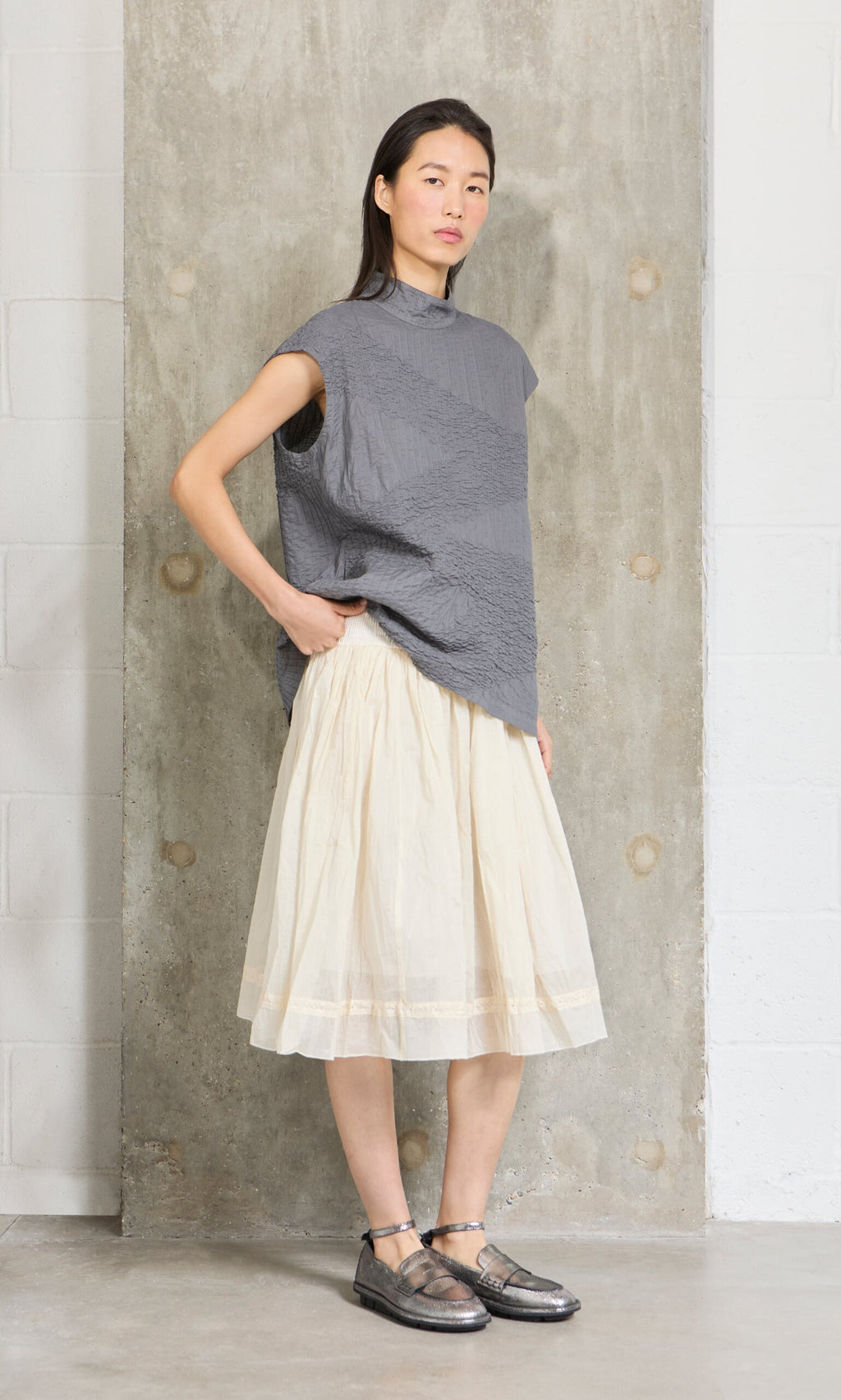 Plümo- Woman in a gray textured top and the Lou Organza Skirt stands against a concrete wall, gazing confidently at the camera.