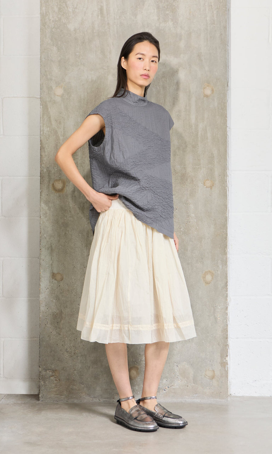 Plümo- Woman in a gray textured top and the Lou Organza Skirt stands against a concrete wall, gazing confidently at the camera.