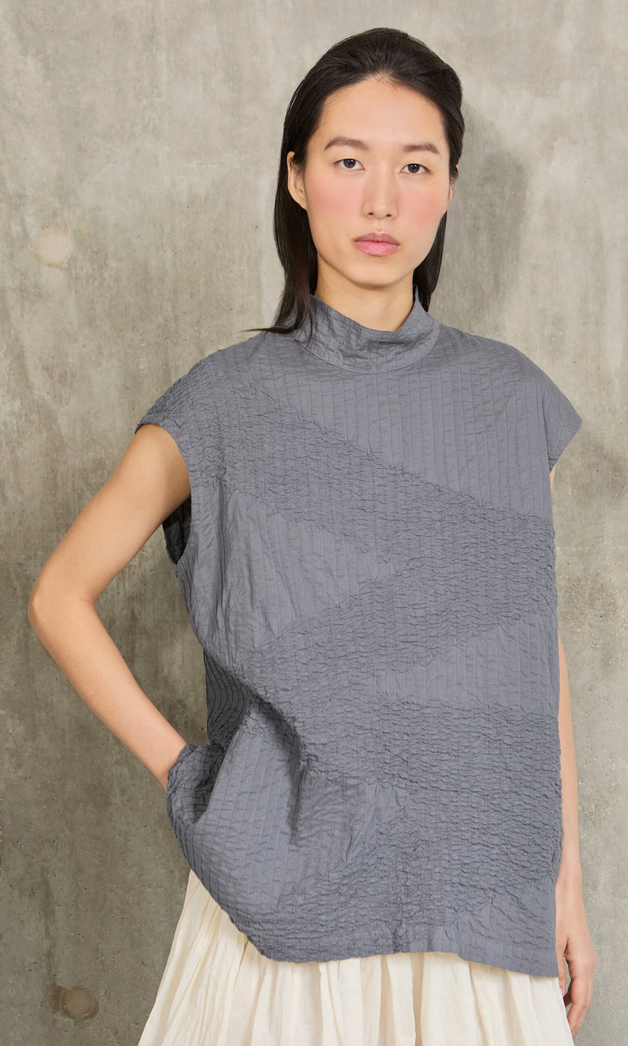 Plümo- Woman with long dark hair wears a Minato Top and cream skirt, posing against a concrete wall.