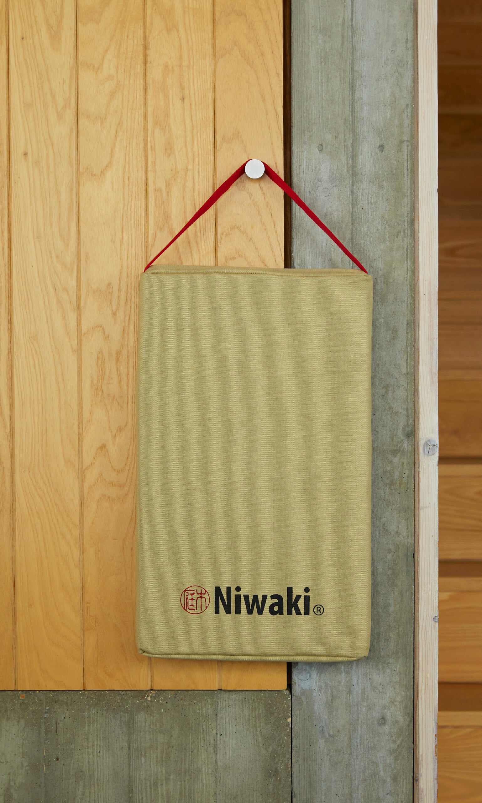 Plümo- A tan Niwaki Kneeler with a red strap hangs by a hook, next to a weatherproof knee cushion.