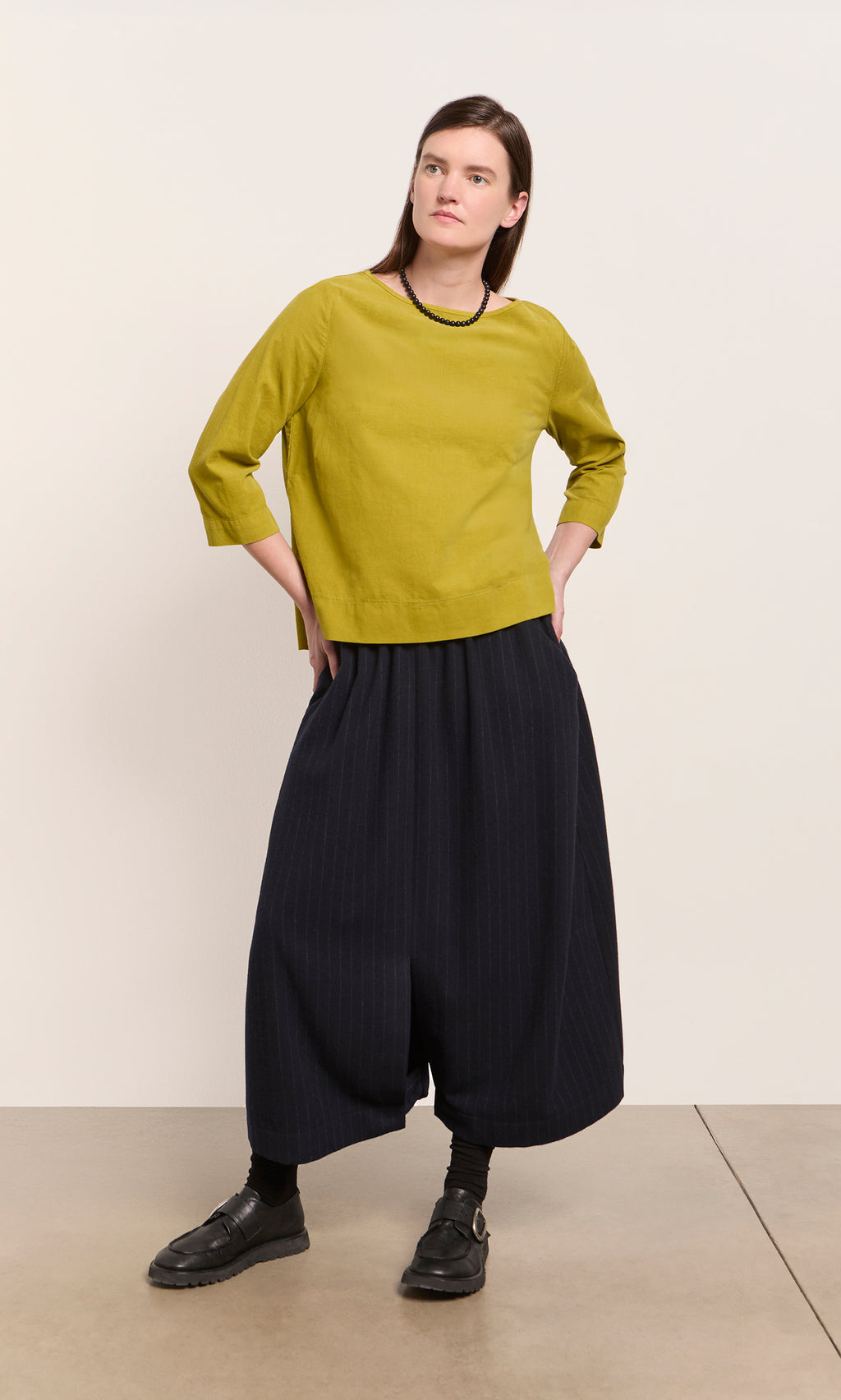 Plümo  Woman in a Moss Top - Corduroy and wide black pants stands with hands on hips against a plain background.