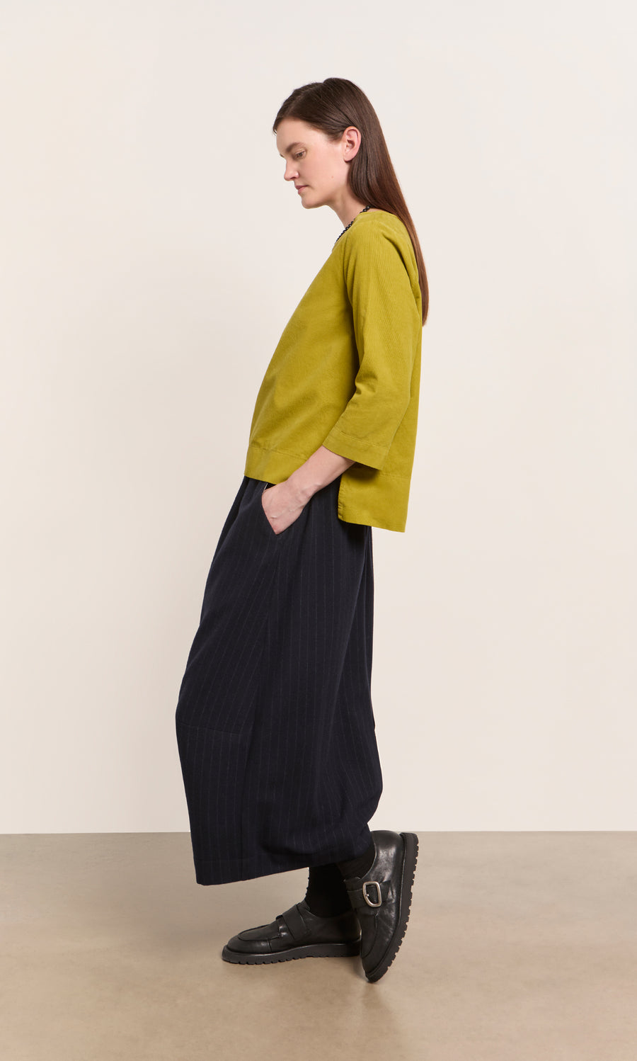 Plümo  Woman in a Moss Top - Corduroy and wide black pants stands sideways, hands in pockets.