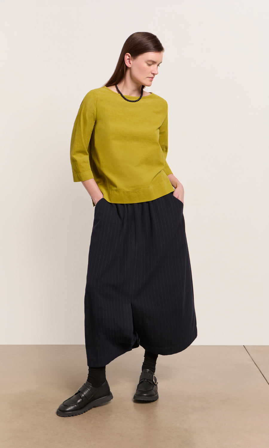 Plümo  Woman in a Moss Top - Corduroy, relaxed skirt, and black shoes stands against a plain background.
