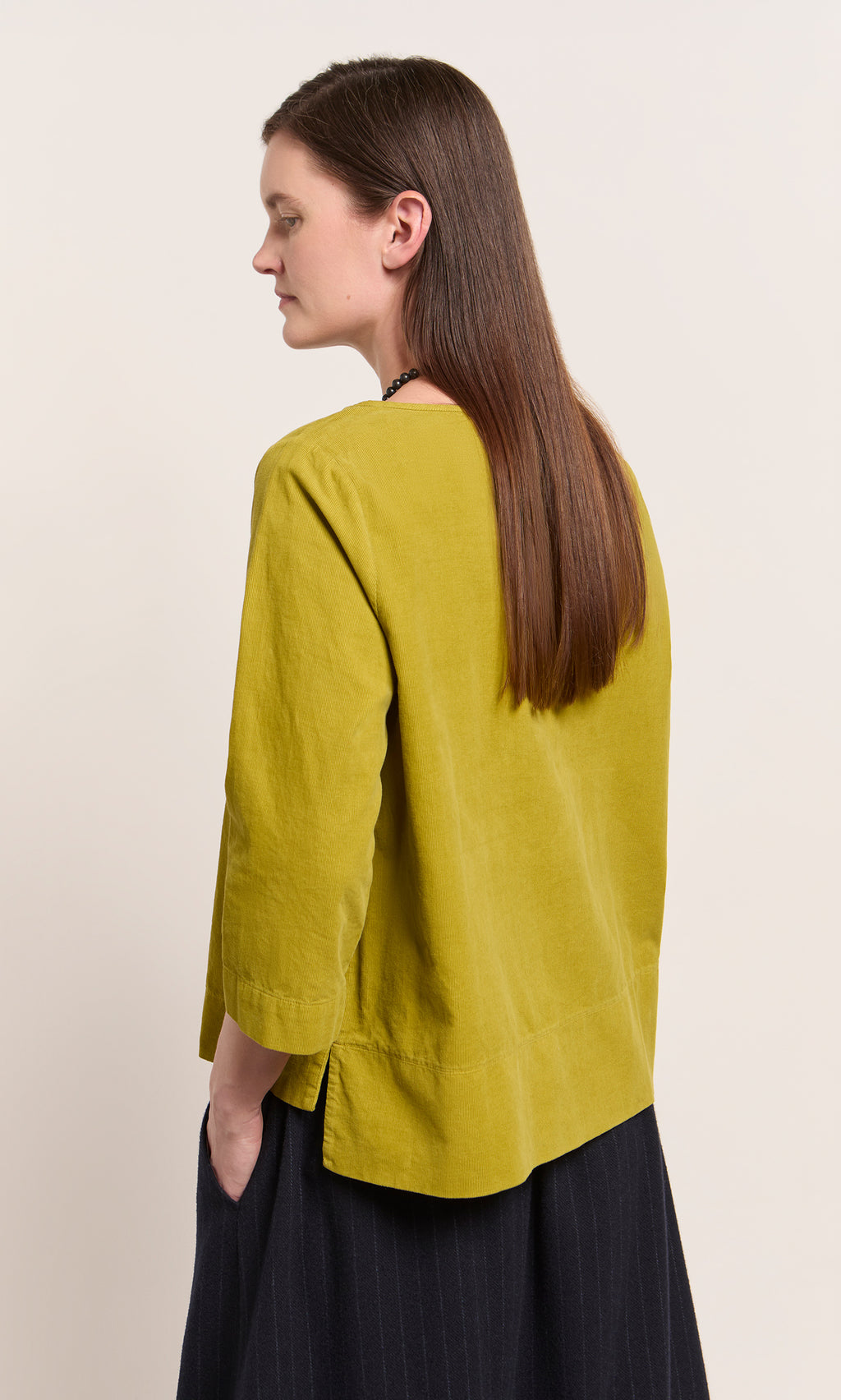 Plümo  Woman with long brown hair in a relaxed fit Moss Top - Corduroy and dark skirt, hands in pockets, side view.