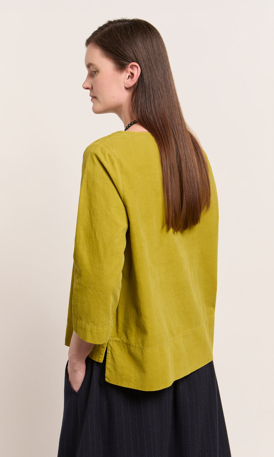 Plümo  Woman with long brown hair in a relaxed fit Moss Top - Corduroy and dark skirt, hands in pockets, side view.
