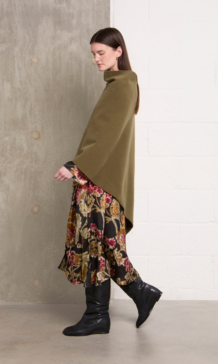 Plümo  Woman in a Moss Poncho with cowl neck, floral skirt, and black boots walks by a white brick wall indoors.