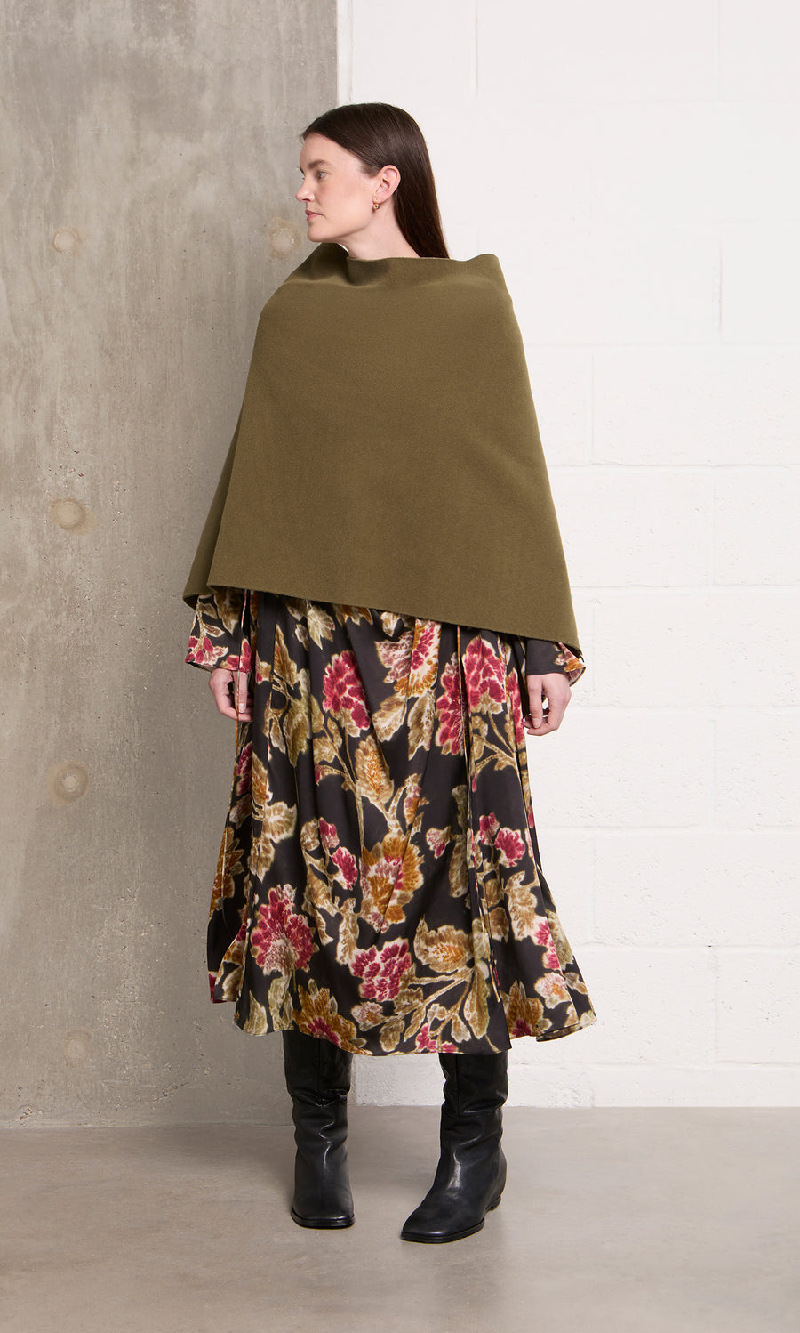 Plümo  Woman wearing the Moss Poncho, floral dress, and black boots stands by a white brick wall, looking to the left.