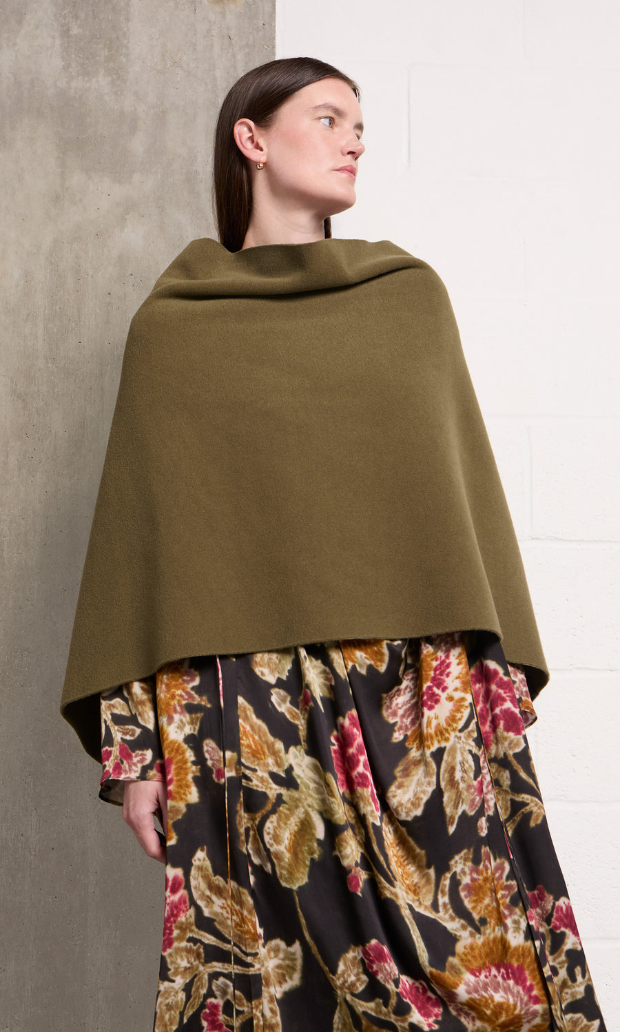 Plümo  Woman in floral dress and Moss Poncho stands by concrete and white brick wall, looking to the side.
