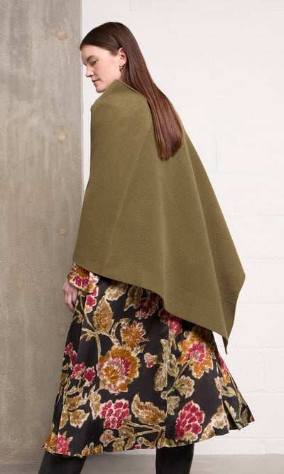 Plümo  Woman in a floral dress and Moss Poncho stands sideways by a concrete wall, looking down.