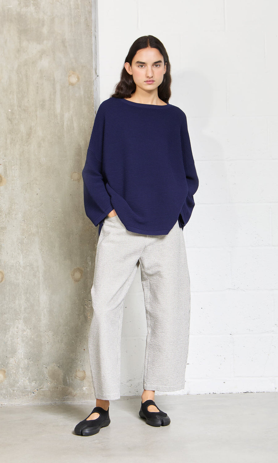 Plümo- Person with long dark hair wears the Joya Sweater - Deep Navy, light striped pants, and black shoes against a concrete wall.