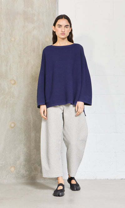 Plümo- Woman wearing Plümo Studio's Joya Sweater in Deep Navy, wide light gray pants, and black shoes by a concrete wall.