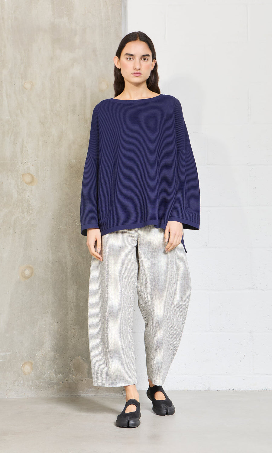 Plümo- Woman wearing Plümo Studio's Joya Sweater in Deep Navy, wide light gray pants, and black shoes by a concrete wall.