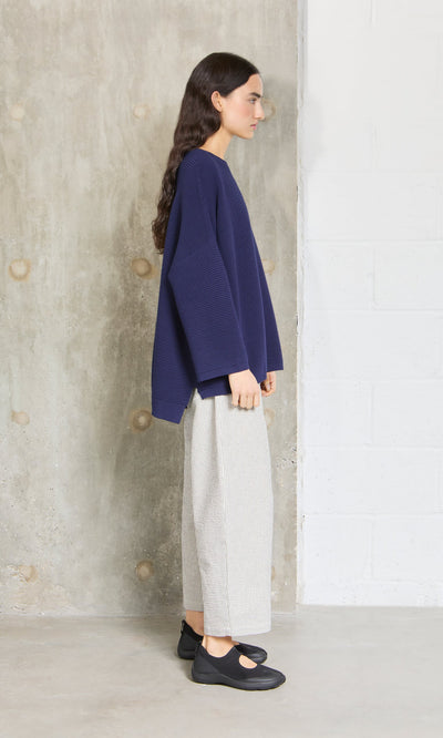 Plümo- Woman in a Joya Sweater - Deep Navy by Plümo Studio, wide-leg light gray pants, and black shoes stands by a concrete wall.