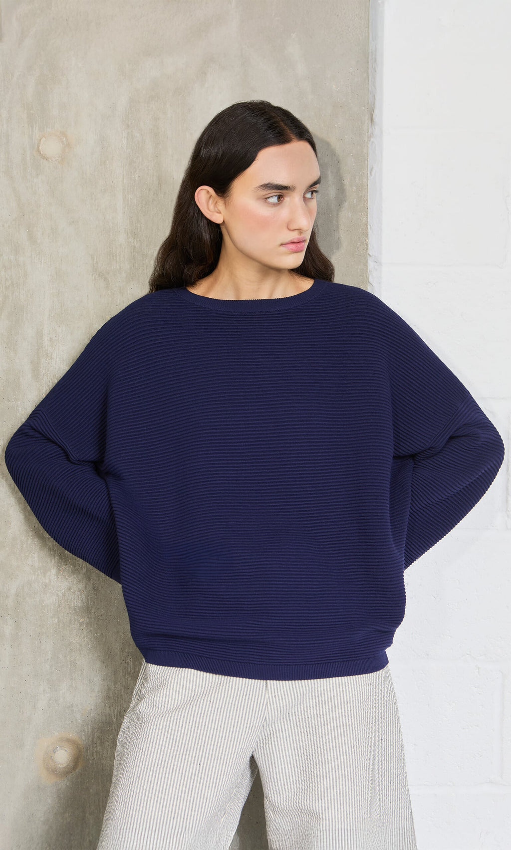 Plümo- Woman with long dark hair wears the Joya Sweater in Deep Navy, standing by a concrete and white wall.