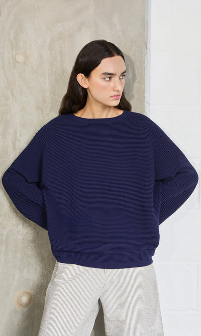 Plümo- Woman with long dark hair wears the Joya Sweater in Deep Navy, standing by a concrete and white wall.