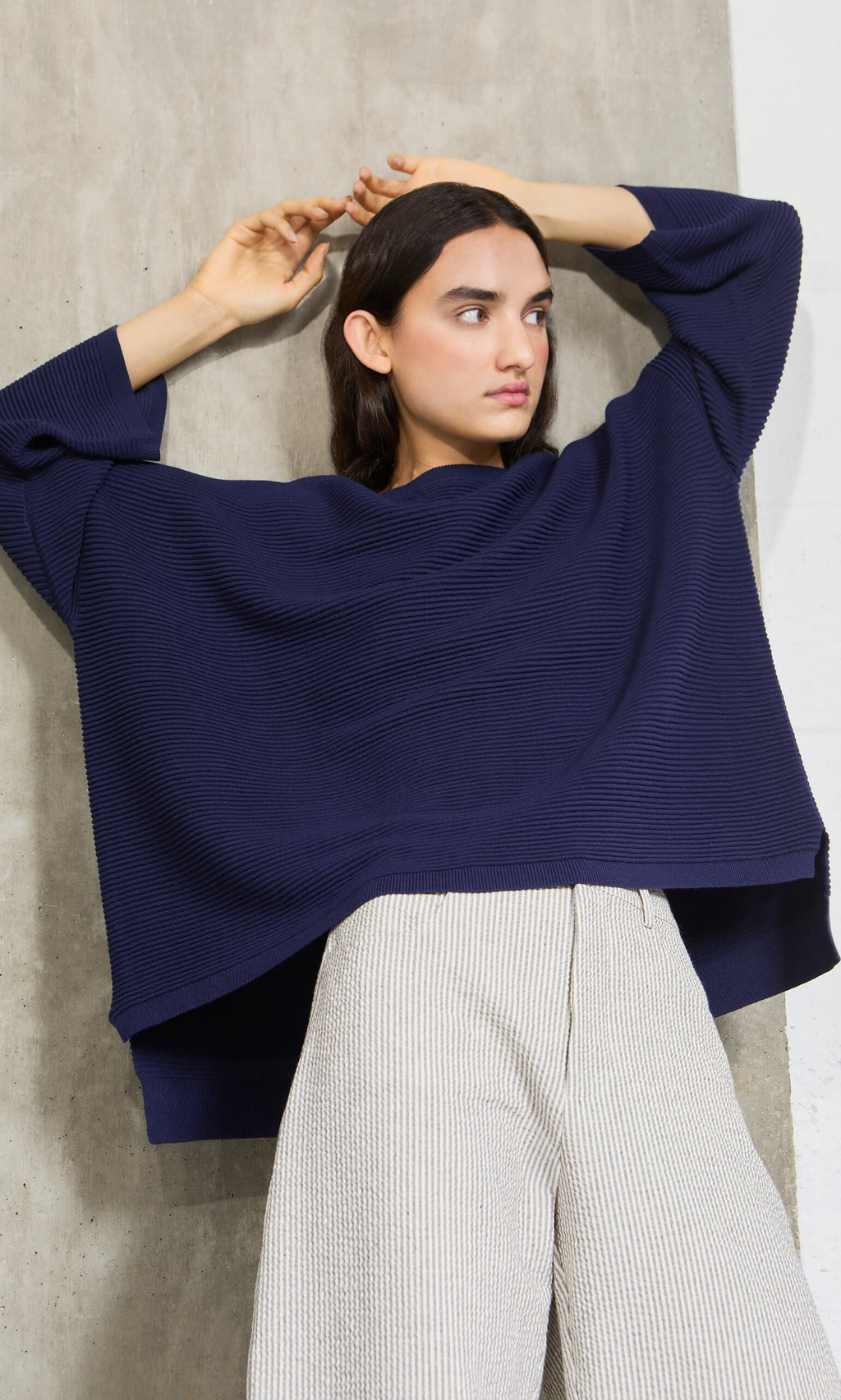 Plümo- Woman wears the Joya Sweater in Deep Navy with light pants, posing with arms raised by a concrete wall.