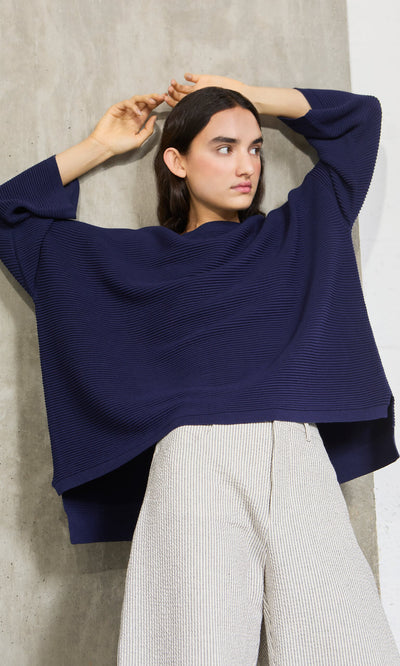 Plümo- Woman wears the Joya Sweater in Deep Navy with light pants, posing with arms raised by a concrete wall.