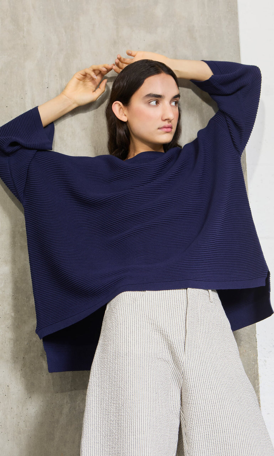Plümo- Woman wears the Joya Sweater in Deep Navy with light pants, posing with arms raised by a concrete wall.