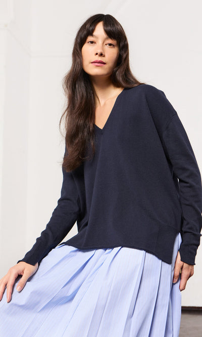 Plümo- Woman with long brown hair wears a Navy V Neck and a light blue pleated skirt, seated indoors.