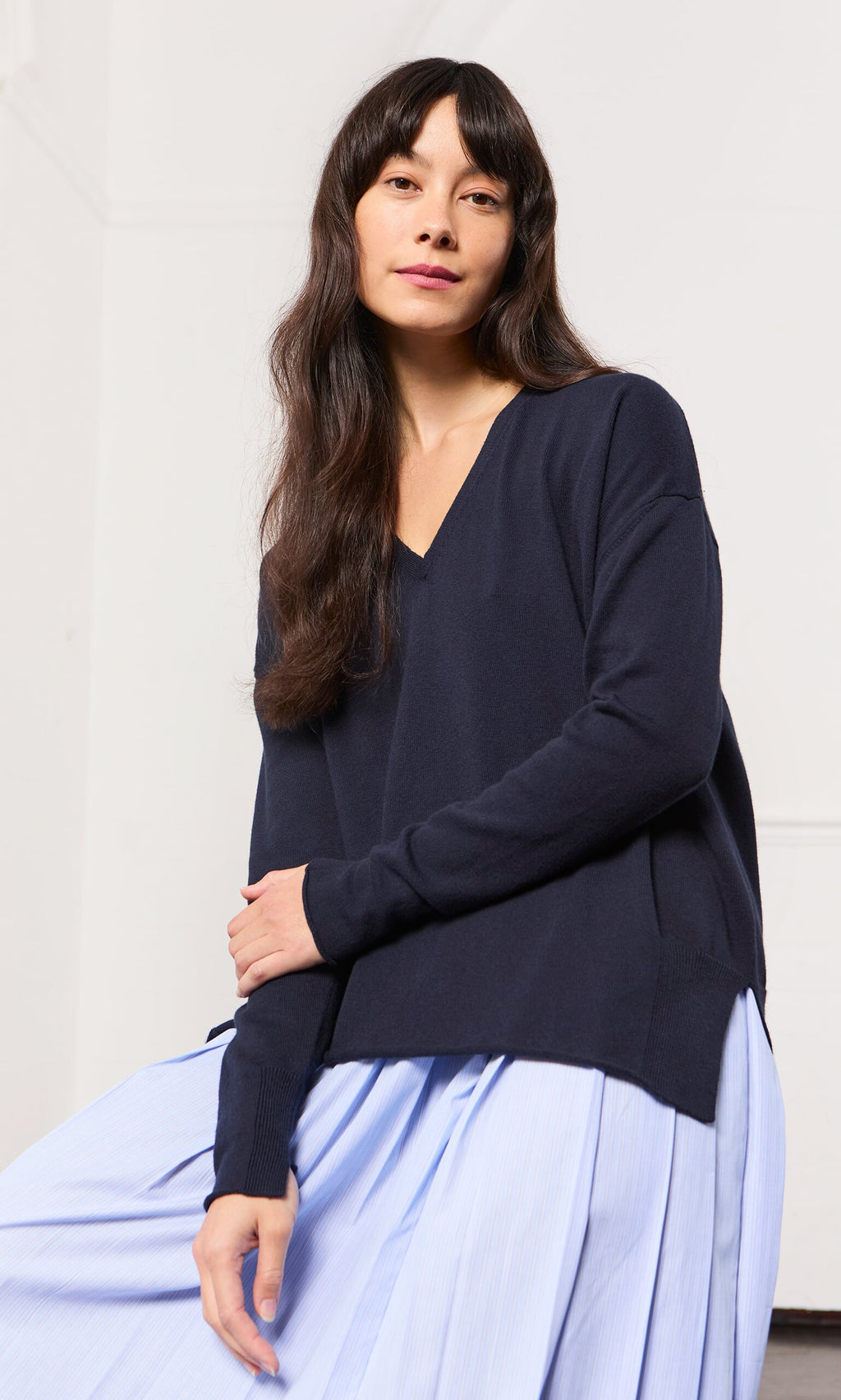 Plümo- Woman with long dark hair in a Navy V Neck sweater and light blue skirt, sitting and looking at the camera.