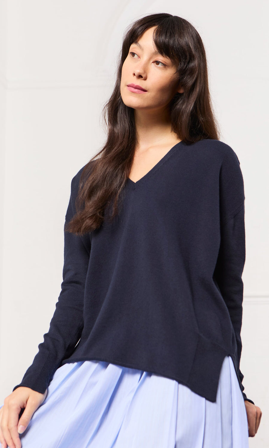 Plümo- Woman wearing a Navy V Neck sweater and a light blue pleated skirt, gazing slightly upward with a neutral expression.