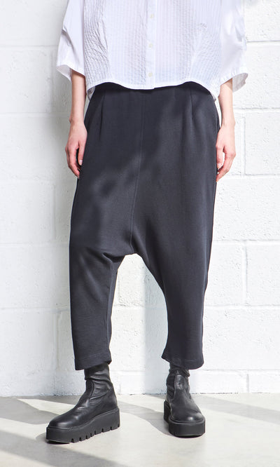 Plümo- Person in black Nerula Pants, cropped legs, black boots, and a white textured shirt by a white wall.