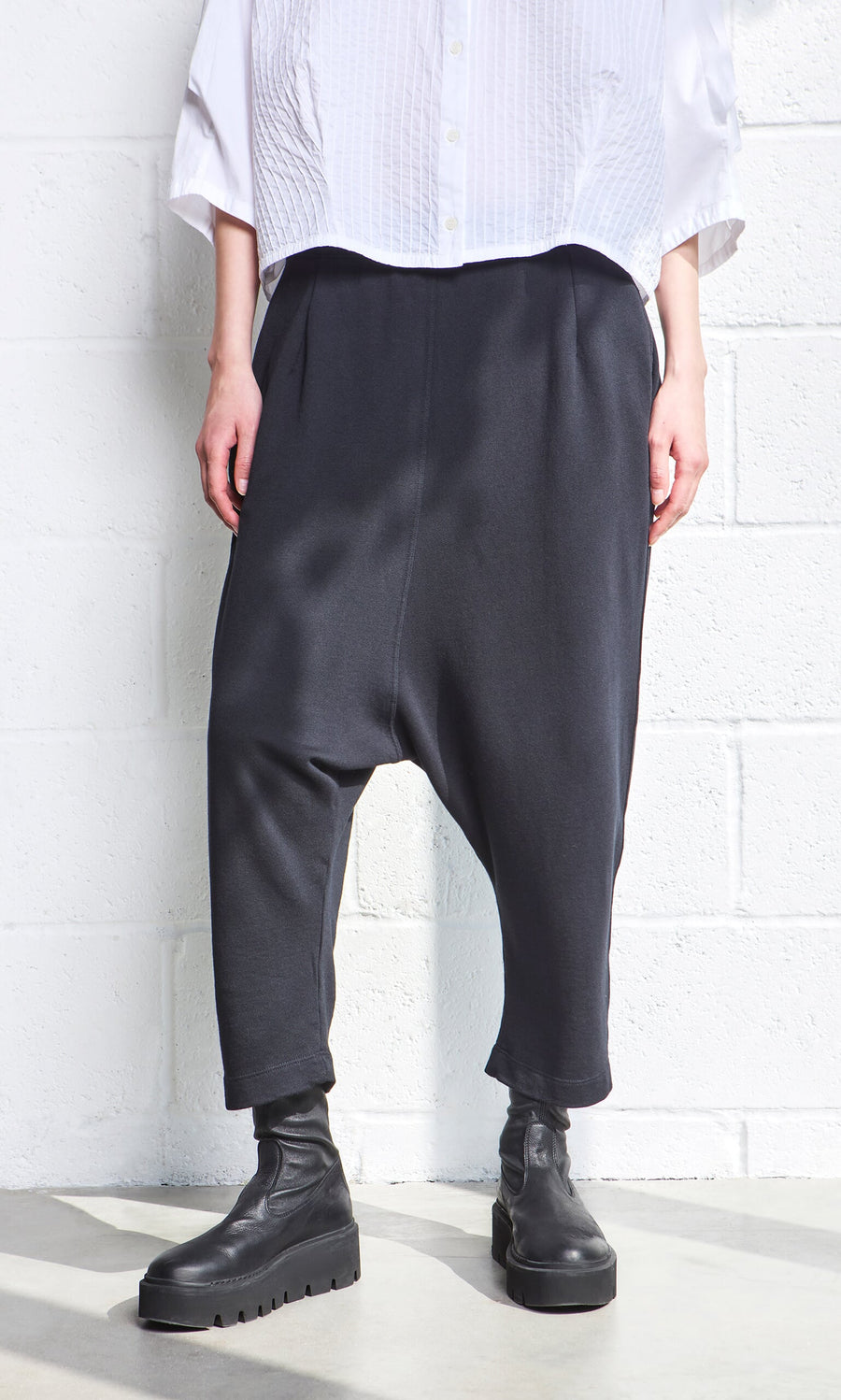 Plümo- Person in black Nerula Pants, cropped legs, black boots, and a white textured shirt by a white wall.