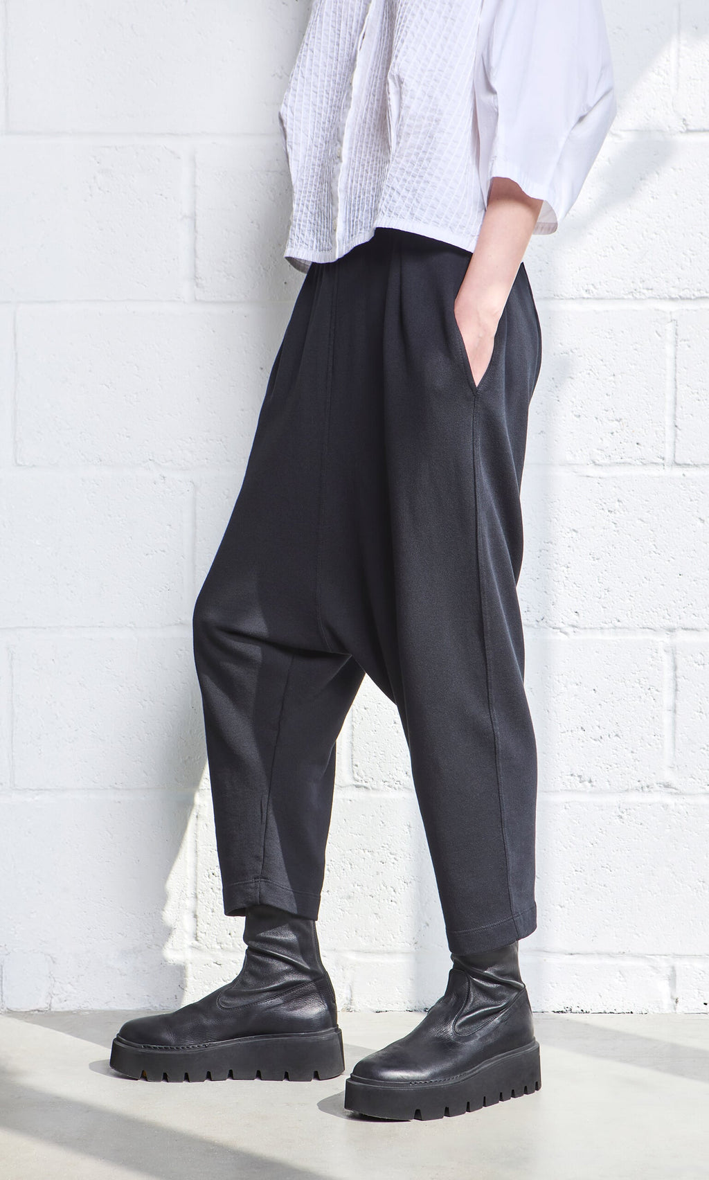 Plümo- Person in Nerula Pants, black boots, and a white textured top stands against a white brick wall.