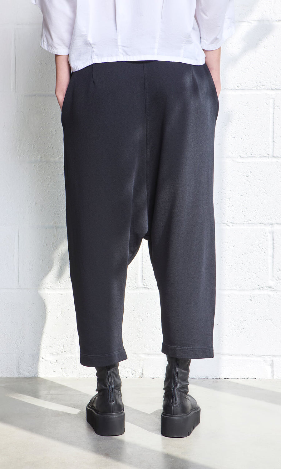 Plümo- Person wearing black Nerula Pants, black platform boots, and a white shirt, standing against a white wall.