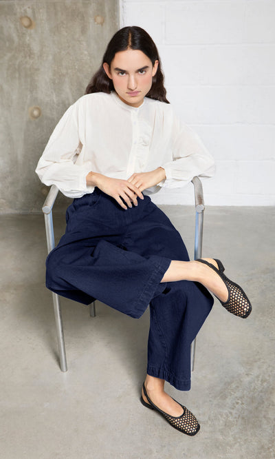 Plümo- Person in a white blouse and navy pants sits on a metal chair, wearing Noa Slingbacks inspired by Danish design.