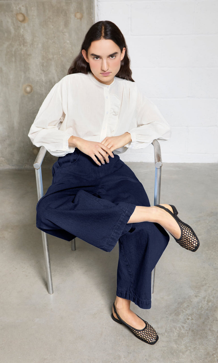 Plümo- Person in a white blouse and navy pants sits on a metal chair, wearing Noa Slingbacks inspired by Danish design.