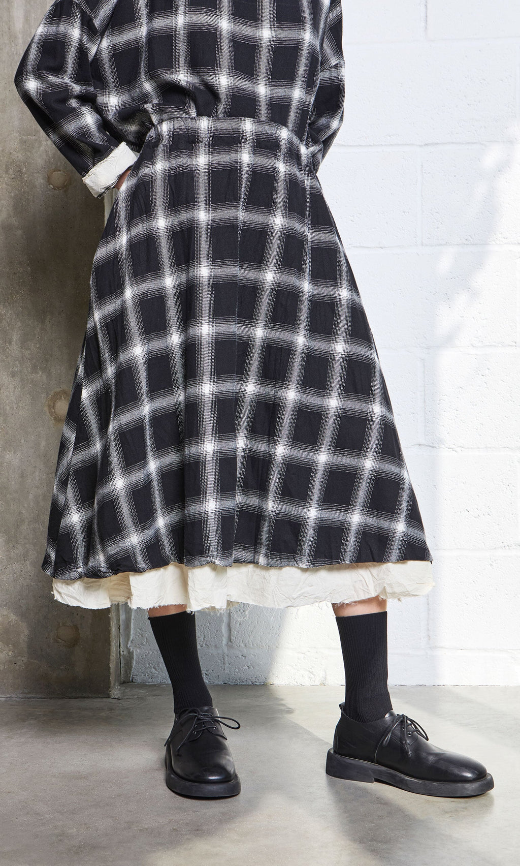 Plümo- Person wearing the Noemi Skirt with an elasticated waistband, black socks and shoes, standing indoors by a concrete wall.