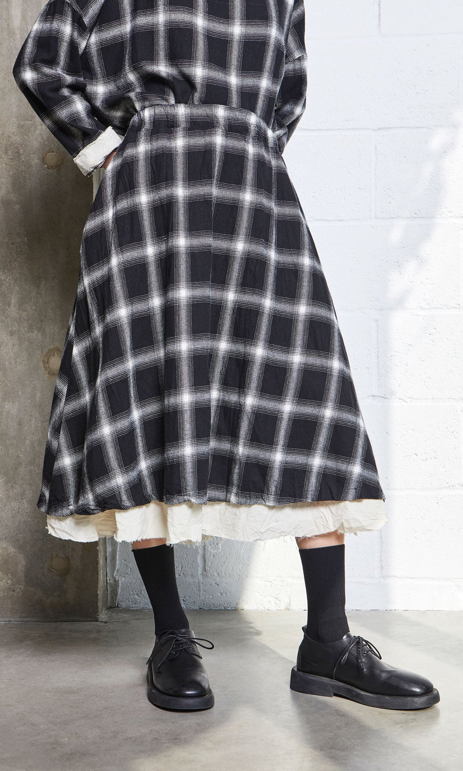 Plümo- Person wearing the Noemi Skirt with an elasticated waistband, black socks and shoes, standing indoors by a concrete wall.