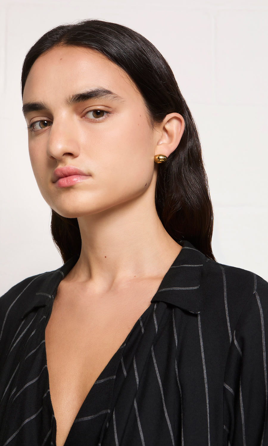 Plümo  Woman with long dark hair wears Oval Studs 14K gold-plated, nickel free and a black pinstripe blouse, facing camera.