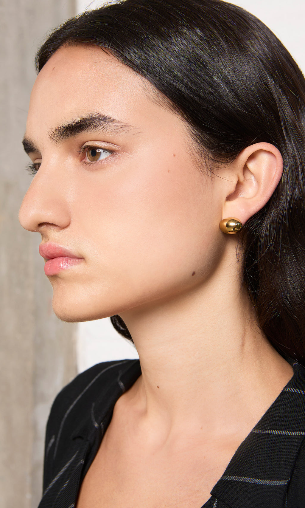 Plümo  Person with long dark hair in a black blazer and Oval Studs, facing left.