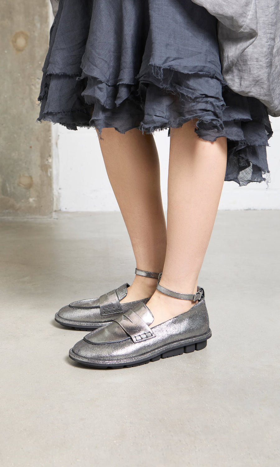 Plümo- Person wearing a dark ruffled skirt and Silver Loafers, standing on a concrete floor.